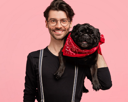 Smiling man with glasses holding a happy pug in a red bandana on a pink background